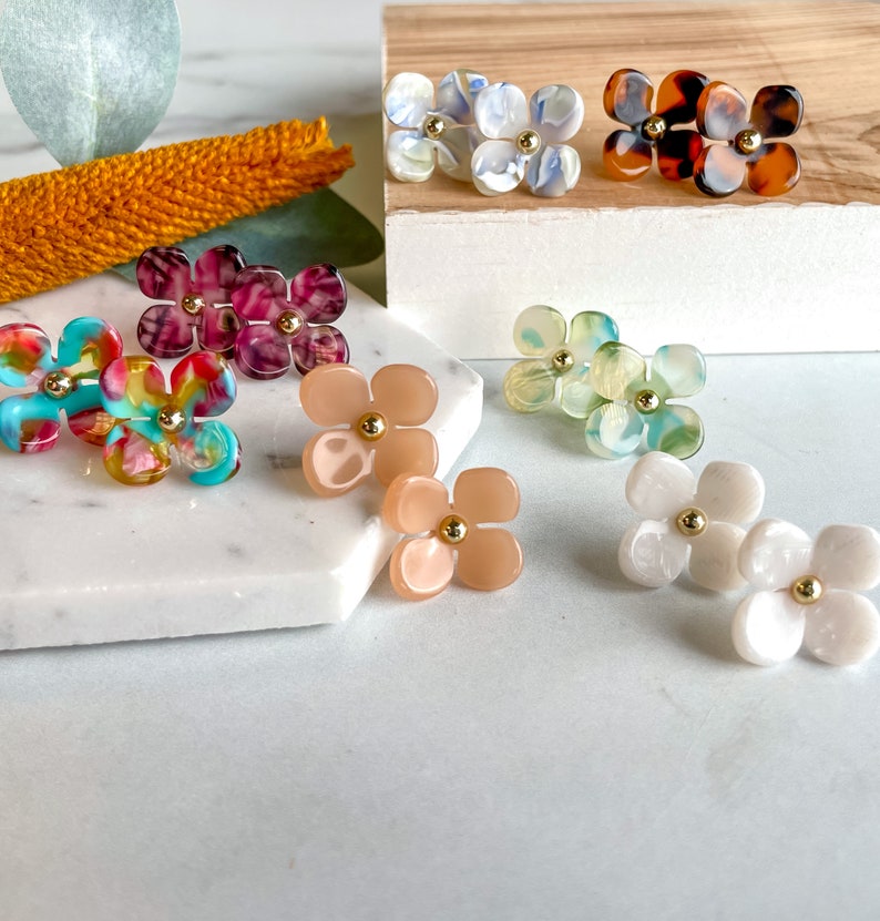 Puede incluir: Surtido de pendientes con forma de flor de colores con detalles dorados. Los pendientes est&aacute;n hechos de varios materiales, incluyendo pl&aacute;stico, resina y concha.