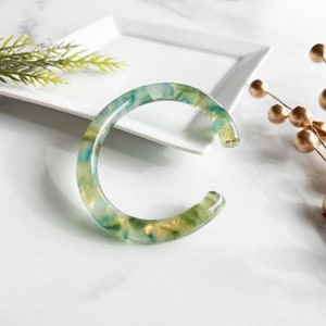 May include: A green and gold marbled bangle bracelet. The bracelet is made of a smooth, glossy material and has a simple, elegant design.