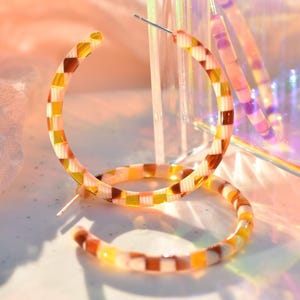May include: A pair of hoop earrings with a checkered pattern in shades of brown, yellow, and white. The earrings are made of a translucent material and have a silver post. The earrings are displayed on a white surface with a soft, colorful background.