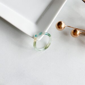 May include: A clear, translucent ring with a swirl of blue and green colors. The ring is made of resin and has a simple, modern design.