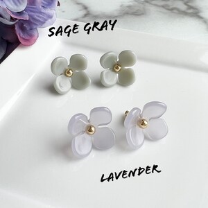 May include: Two pairs of flower-shaped earrings. The first pair is sage grey with a gold centre. The second pair is lavender with a gold centre. The text "Sage Gray" and "Lavender" is written below each pair of earrings.