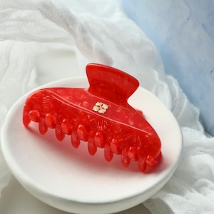 May include: A vibrant red hair claw clip with a textured surface and a gold-colored accent. The claw clip is resting on a white ceramic dish, with a soft white fabric in the background. This hair accessory is a stylish addition to any hairstyle.