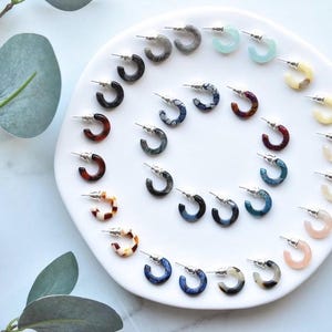 May include: A collection of small hoop earrings arranged in a spiral pattern on a white circular plate. The earrings feature various colors and patterns, including tortoiseshell, solid pastels, marbled blues, greys, and blacks, all with silver posts.