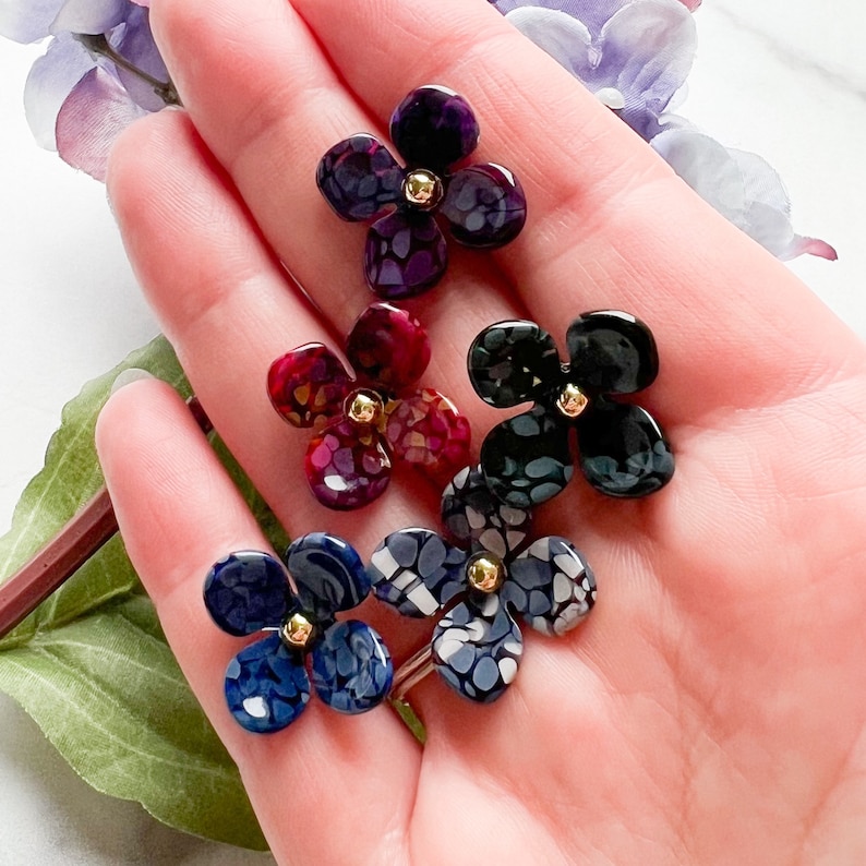 May include: Five resin flower charms in various colours and patterns, including purple, red, black, and white. Each charm has a gold centre.