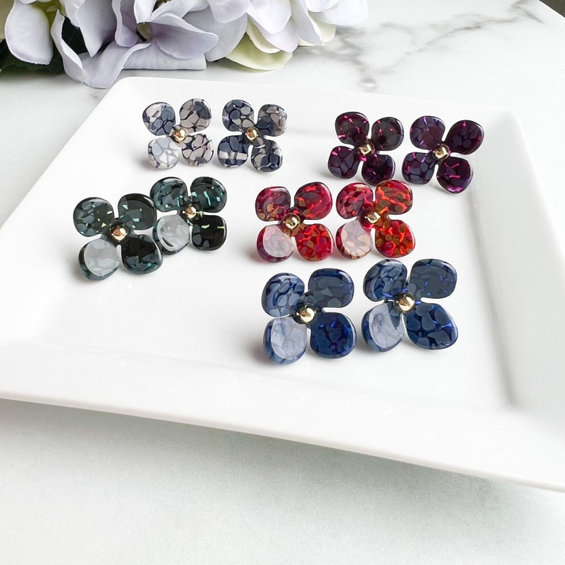 May include: Assortment of colourful flower-shaped earrings with a marbled pattern. The earrings are in shades of blue, green, red, and grey.