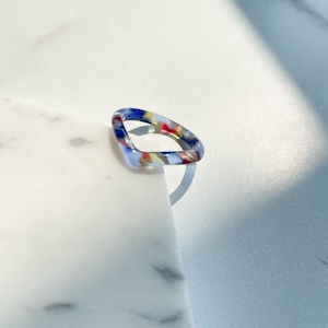 May include: A colorful, geometric ring made of resin. The ring is a square shape with rounded edges and features a blue, red, yellow, and white pattern.