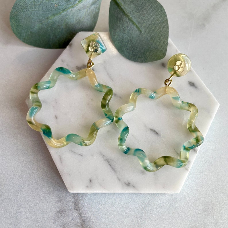 Puede incluir: Un par de pendientes de aro con estampado de espiral verde y amarillo con detalles dorados. Los pendientes tienen forma de estrella con bordes redondeados.