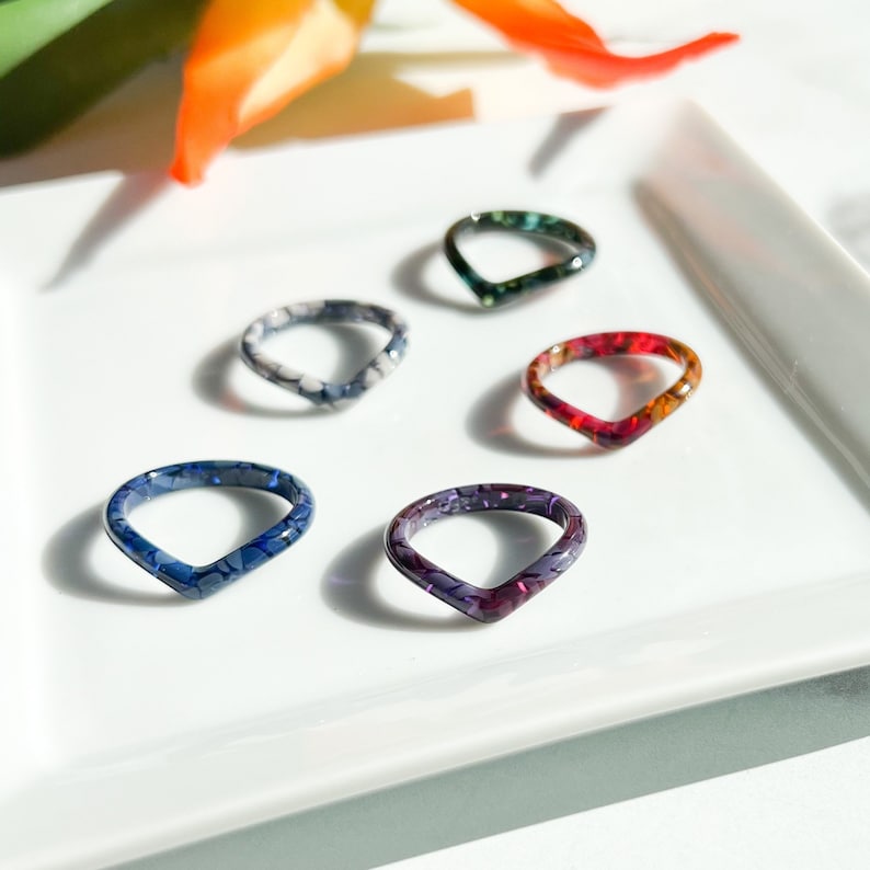 May include: Six colorful, v-shaped rings made of resin with a marbled pattern. The rings are arranged on a white plate.