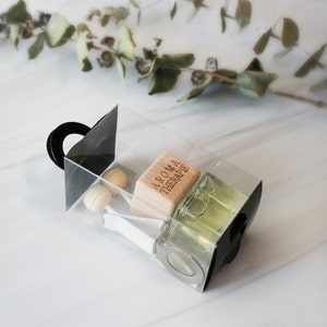 Puede incluir: Una caja transparente con asa negra contiene una pequeña botella de vidrio con líquido verde claro, un bloque de madera con "AROMA THERAPIE" impreso y dos pequeñas esferas de madera. La caja está sobre una superficie blanca.