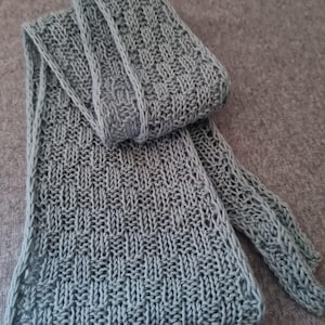 May include: A light blue knitted scarf with a textured pattern. The scarf is folded, showcasing the intricate stitch work. The ends of the scarf are visible, displaying a delicate openwork design. The scarf is made of soft yarn.