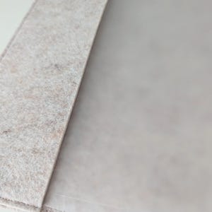 May include: A light beige felt folder featuring a textured, fibrous surface with a soft, matte appearance. The folder has a clean, straight edge and a smooth, semi-transparent interior panel that is partially visible, showing a subtle, neutral tone.