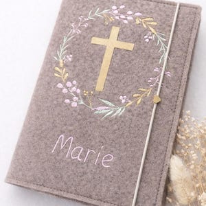 Könnte beinhalten: Ein grauer Filz-Buchumschlag mit einem goldenen Kreuz und floralen Stickereien. Der Umschlag trägt den Namen "Marie" in zarter Schrift gestickt. Ein weißes Gummiband mit einem kleinen goldenen Herz verschließt den Umschlag.