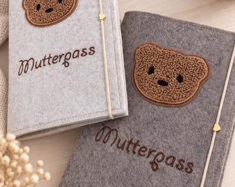 May include: Two rectangular felt covers for a Mutterpass, one in light gray and one in dark gray. Each features a brown textured bear face patch and the embroidered text Mutterpass. A thin elastic band with a small gold heart charm secures each.