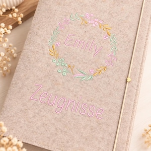 May include: A light beige felt notebook cover featuring an embroidered floral wreath in pastel pink, green, and yellow tones. The name Emily is stitched in pink inside the wreath, with the word Zeugnisse embroidered in pink below the wreath.