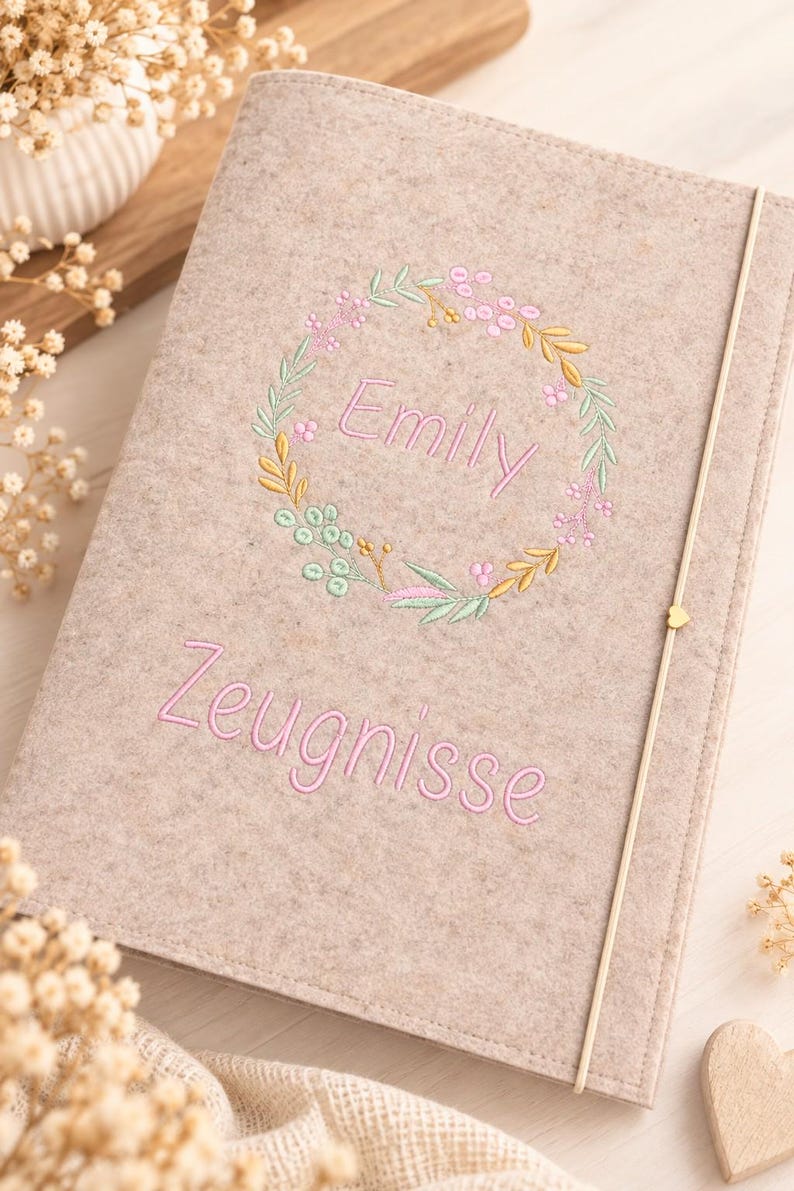 May include: A light beige felt notebook cover featuring an embroidered floral wreath in pastel pink, green, and yellow tones. The name Emily is stitched in pink inside the wreath, with the word Zeugnisse embroidered in pink below the wreath.