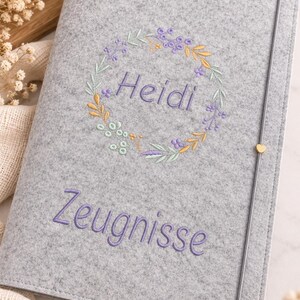 May include: A light gray felt document folder featuring an embroidered floral wreath surrounding the name Heidi in purple thread. Below the wreath, the word Zeugnisse is embroidered in purple. A thin gray elastic band with a small gold heart.