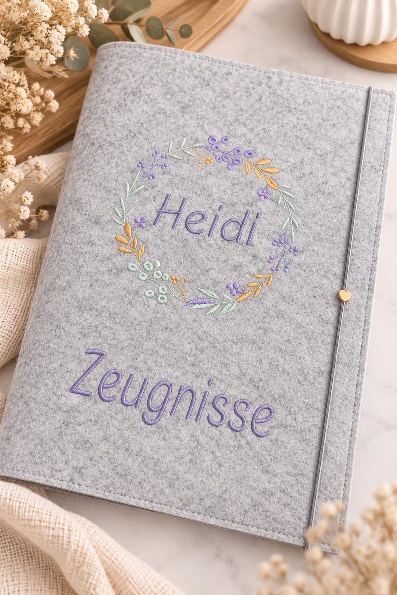 May include: A light gray felt document folder featuring an embroidered floral wreath surrounding the name Heidi in purple thread. Below the wreath, the word Zeugnisse is embroidered in purple. A thin gray elastic band with a small gold heart.