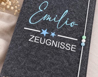 Könnte beinhalten: Eine dunkelgraue Filzmappe mit dem Namen Emilio in hellblauer Schreibschrift, zwei hellblauen Sternen und dem Wort ZEUGNISSE in weißen Großbuchstaben. Ein weißes Gummiband mit drei kleinen Perlen in Grün, Weiß und Blau hält die Mappe zusammen.