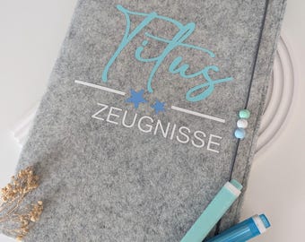 Könnte beinhalten: Ein grauer Filz-Dokumentenhalter mit dem Namen "Titus" in hellblauer Schrift und dem Wort "ZEUGNISSE" in weißen Blockbuchstaben. Zwei blaue Textmarker und eine dekorative Schnur mit Perlen sind ebenfalls zu sehen.