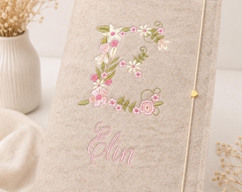 May include: A beige felt notebook cover featuring an embroidered floral letter E in shades of pink, white, and green. Below the letter, the name Elin is embroidered in pink script. A thin elastic band with a small gold heart charm secures the cover.