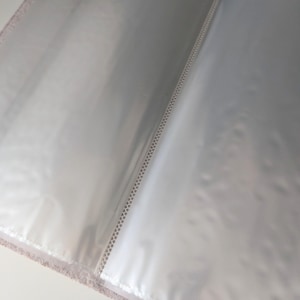 May include: An open photo album featuring clear plastic sleeves for document or photograph storage. The album has a textured, light-colored cover visible along the edges and a central spine with a white stitched binding detail holding the pages.