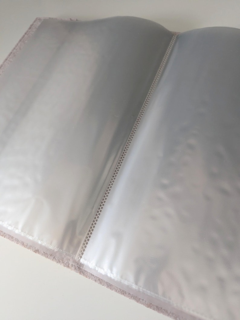 May include: An open photo album featuring clear plastic sleeves for document or photograph storage. The album has a textured, light-colored cover visible along the edges and a central spine with a white stitched binding detail holding the pages.