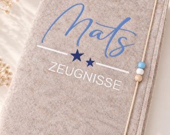 Könnte beinhalten: Eine hellbeige Filzmappe mit dem in Blau geschriebenen Namen Mats, zwei dunkelblauen Sternen und dem weißen Schriftzug ZEUGNISSE. Ein Gummiband mit einer hellblauen, einer weißen und einer Holzperle hält die Mappe sicher verschlossen.