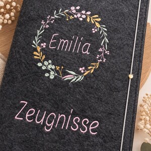May include: A dark gray felt folder featuring an embroidered floral wreath surrounding the name Emilia in pink thread. Below the wreath, the word Zeugnisse is embroidered in pink. A thin vertical elastic band with a small heart charm secures it.