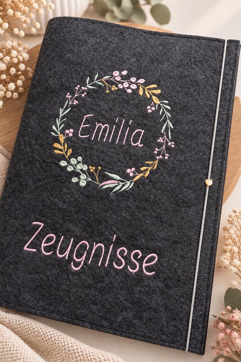 May include: A dark gray felt folder featuring an embroidered floral wreath surrounding the name Emilia in pink thread. Below the wreath, the word Zeugnisse is embroidered in pink. A thin vertical elastic band with a small heart charm secures it.