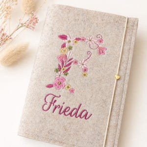 Könnte beinhalten: Ein hellbeiger Notizbuchumschlag aus Filz mit einem gestickten floralen Buchstaben F in Rosa-, Gelb- und Grüntönen. Unter dem Buchstaben ist der Name Frieda in dunkelrosa Schreibschrift gestickt. Ein dünnes Gummiband mit einem kleinen goldenen Herz.
