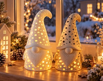 May include: Two white ceramic gnome figurines with pointed hats and long beards, featuring intricate circular cutouts that allow warm light to glow from within. The gnomes are positioned side by side, displaying a smooth matte finish and hollow bodies.