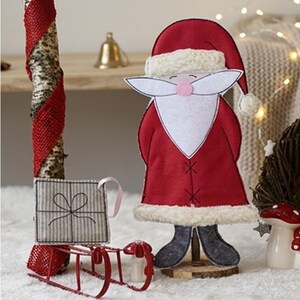 Pode incluir: Um conjunto de 5 peças com uma figura de Pai Natal em tecido com casaco vermelho e rebordo branco, uma pequena caixa de presente às riscas cinzentas e um trenó de metal vermelho. O texto diz 5 tlg. Set, Weihnachtsmann e ITH-Stickdatei.