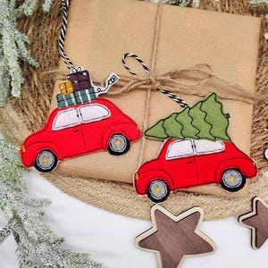 Op de afbeelding: Twee geborduurde viltornamenten in de vorm van rode auto's. De ene auto draagt een stapel kleurrijke cadeautjes op het dak, terwijl de andere een groene kerstboom draagt. Beide ornamenten hebben zwart-witte koordjes om op te hangen.