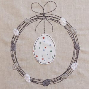 May include: Embroidered Easter wreath with an egg-shaped ornament. The wreath is made of dark grey threads and adorned with embroidered, colourful, oval-shaped accents. The egg-shaped ornament is white with colourful polka dots.