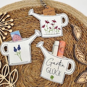 May include: A set of three white watering can-shaped embroidered gift card holders with black outlines. One has blue flowers, another has red flowers, and the third has the words "Garten Glück". The image also includes wooden leaf cutouts.