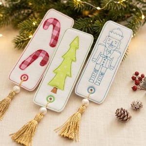 Op de afbeelding: Set van vijf kerstthema bladwijzers. Elke bladwijzer heeft een geborduurd ontwerp: zuurstokken, een kerstboom en een notenkraker. Ze hebben gouden kwasten en parelaccenten. De tekst "5 tlg. Set" is zichtbaar.