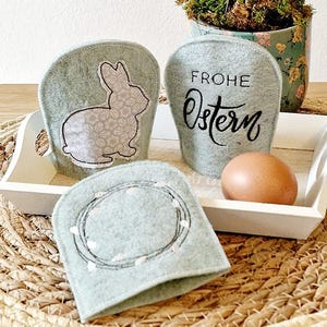 Puede incluir: Un conjunto de cuatro decoraciones de Pascua de fieltro verde claro. Una tiene un diseño de conejito, otra dice "FROHE Ostern", y una tercera tiene un diseño de corona. El conjunto se muestra en una bandeja blanca con un huevo.
