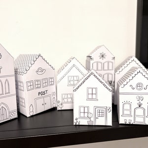 Puede incluir: Una colección de casas de papel blanco con detalles de contorno negro. Las casas varían en diseño, incluyendo una iglesia, una oficina de correos con la palabra "POST" y casas con números. Las casas están dispuestas sobre una estantería negra.