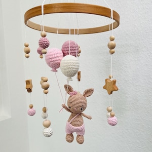 Pode incluir: Um móbile de bebé de madeira com um coelho de crochê rosa e branco segurando balões rosa e branco. O móbile também tem contas e estrelas de madeira.