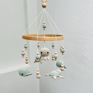 Op de afbeelding: Een handgemaakte baby mobile met gehaakte zeedieren, waaronder een octopus, schildpadden en een walvis, in groen- en beigetinten. De mobile is opgehangen aan een houten ring en versierd met houten kralen, wat een charmante decoratie voor de kinderkamer creëert.