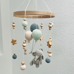 Könnte beinhalten: Ein Holz-Babymobil mit einem gehäkelten Elefanten, Sternen und Ballons in Grün-, Beige- und Brauntönen.