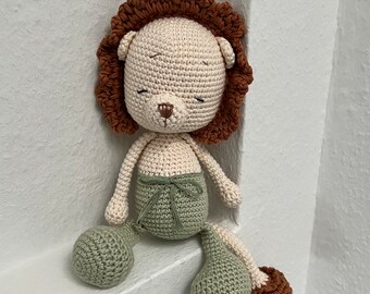 May include: A crocheted lion toy with a brown mane, green pants, and brown feet. The lion is sitting on a white surface.