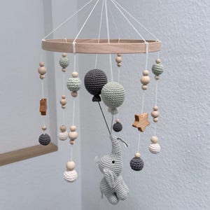 Puede incluir: Móvil de bebé de madera con un marco circular, con elementos de ganchillo colgantes. El móvil incluye bolas de ganchillo grises y blancas, cuentas de madera y un elefante de ganchillo suspendido debajo de globos grises. También se incluye una estrella y un rectángulo de madera.