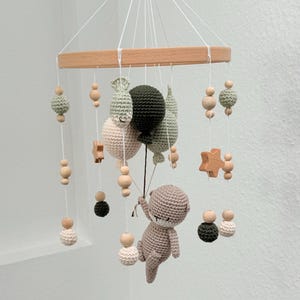 May include: A wooden baby mobile with a crocheted brown bear holding green and cream balloons. The mobile also features wooden beads and stars.