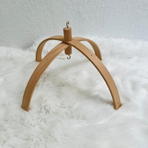 Può includere: Supporto per mobile in legno con finitura naturale. Il supporto ha una base circolare e tre bracci curvi che si estendono verso l'alto. Un gancio è attaccato alla parte superiore del supporto.