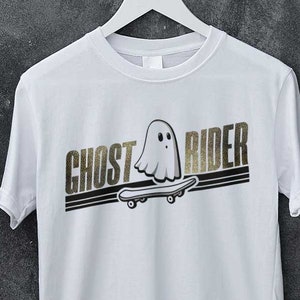 Könnte beinhalten: Weißes T-Shirt mit einer schwarzen und goldenen Grafik eines Geists, der auf einem Skateboard fährt. Der Text "Ghost Rider" ist in schwarzen und goldenen Buchstaben gedruckt.