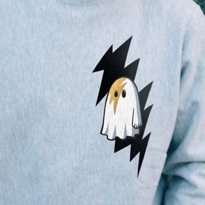 May include: A light gray sweatshirt with a black and white lightning bolt graphic and a white ghost with a yellow face inside the bolt. The text "GEISTES BLITZ" is printed on the shoulder of the sweatshirt.