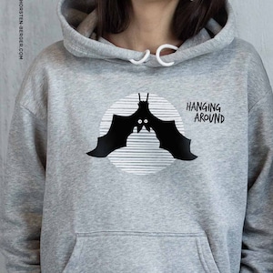 Peut inclure: Un sweat à capuche gris avec un motif de chauve-souris noir et le texte "HANGING AROUND" imprimé sur le devant.