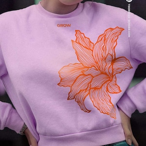 Puede incluir: Una sudadera de color lavanda con un diseño de flor naranja estilizado y la palabra "GROW" impresa en la parte delantera.