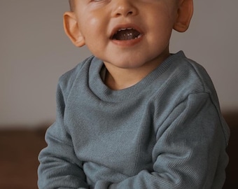 May include: A young child wearing a blue long-sleeved shirt with a small red label that says "Little & Large". The child is looking up and smiling.